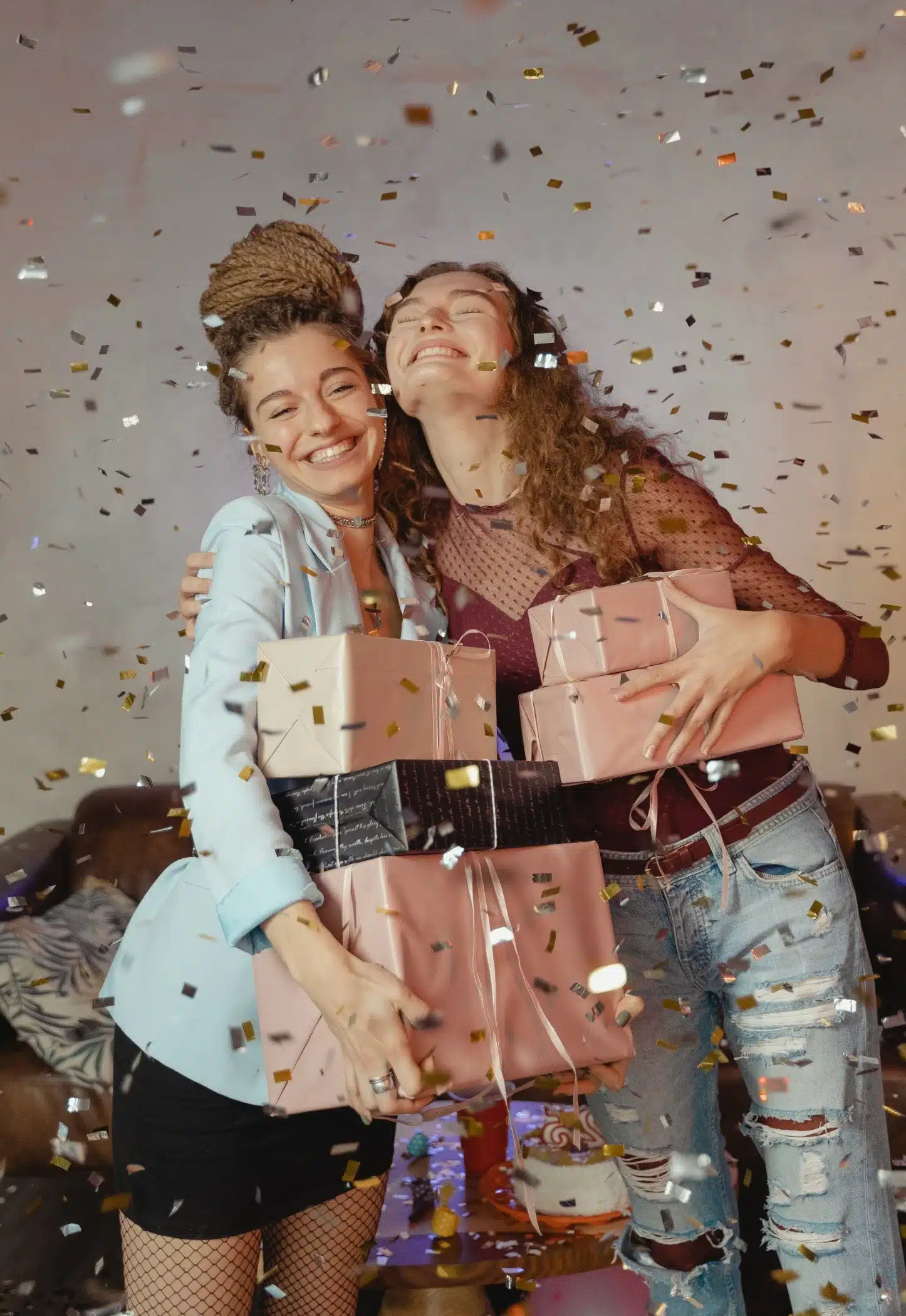 Two sisters with bright smiles, embracing each other in celebration, surrounded by flying confetti and holding pastel-colored gift boxes.