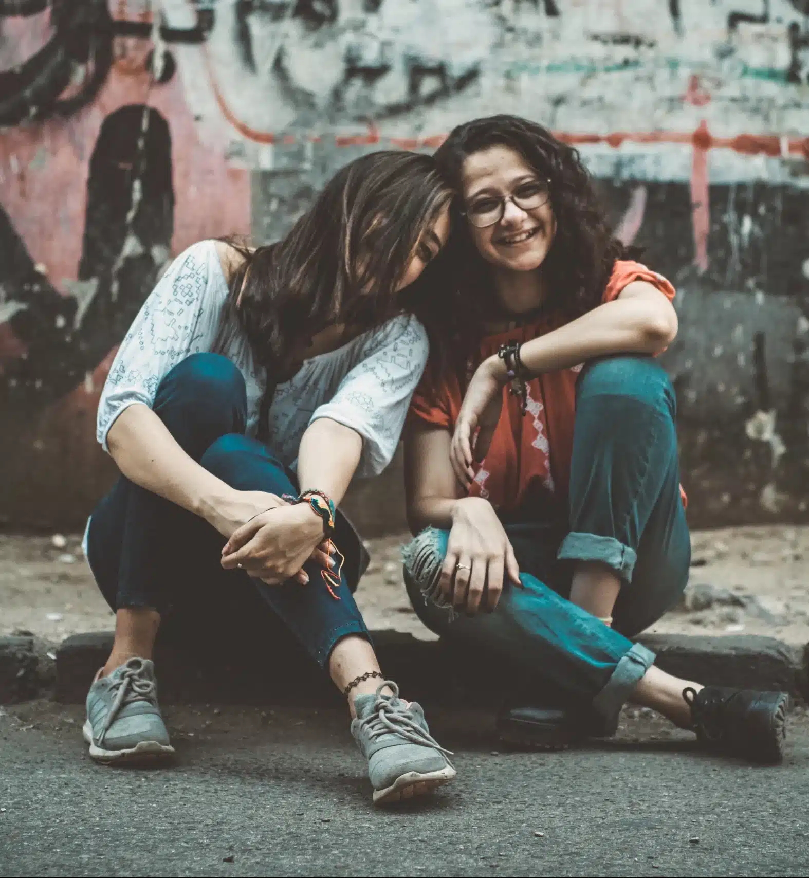 Two happy and smiling sisters sitting together on urban ground and sharing an affectionate moment against a graffiti background.